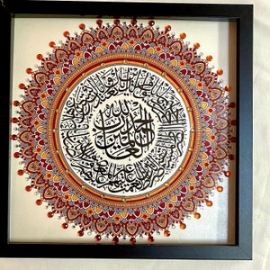 May include: A framed artwork featuring a circular mandala design with intricate details in shades of red, orange, and gold. The center of the mandala contains Arabic calligraphy in black ink.