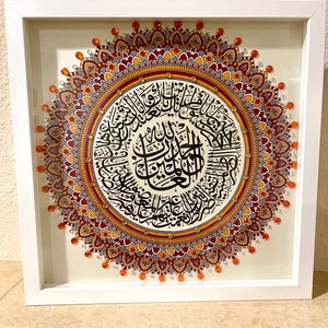 May include: A framed artwork featuring a mandala design with intricate details in shades of orange, red, and purple. The center of the mandala contains Arabic calligraphy in black ink.