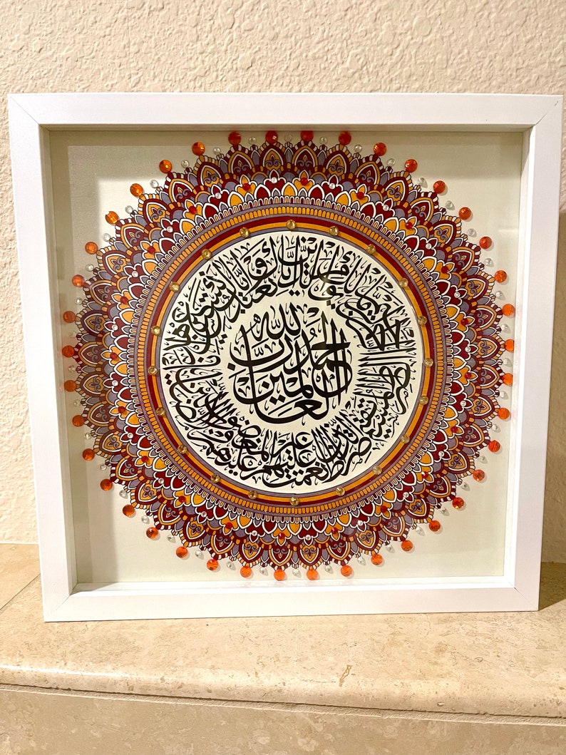 May include: A framed artwork featuring a mandala design with intricate details in shades of orange, red, and purple. The center of the mandala contains Arabic calligraphy in black ink.