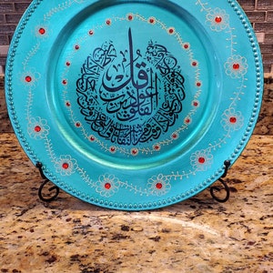 May include: A turquoise charger plate with gold and white floral designs and red rhinestones. The center of the plate features a black Arabic calligraphy design.