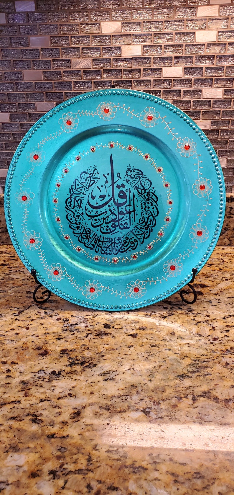 May include: A turquoise charger plate with gold and white floral designs and red rhinestones. The center of the plate features a black Arabic calligraphy design.