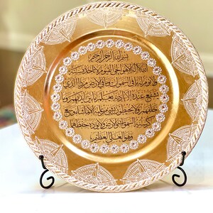 May include: A gold charger plate with a white decorative border and Arabic calligraphy in black. The plate is on a black stand.