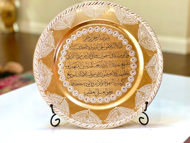 May include: A gold charger plate with a white decorative border and Arabic calligraphy in black. The plate is on a black stand.