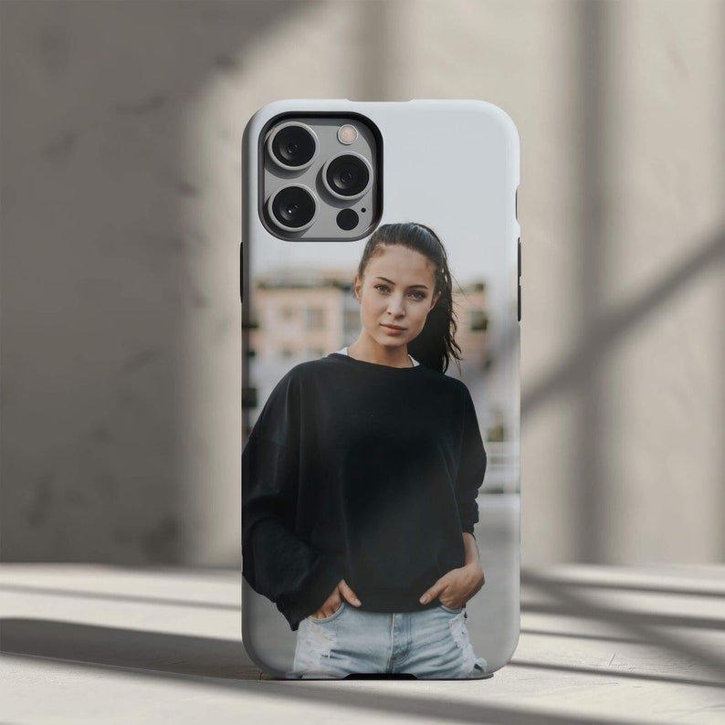 May include: A phone case with a photo of a woman wearing a black sweater and blue jeans. The woman is looking at the camera and has her hair pulled back.