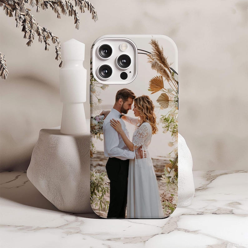 May include: A white phone case with a photo of a couple in wedding attire standing in front of a floral archway.