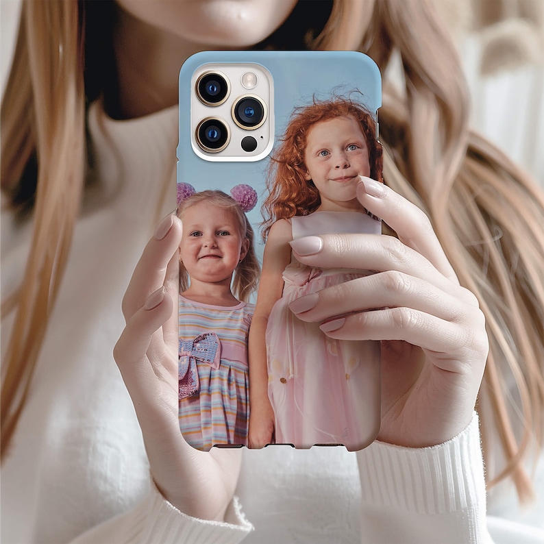May include: A phone case with a photo of two young girls. The girls are wearing dresses and smiling. The case is white with a blue background.
