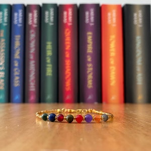 Può includere: Un braccialetto dorato con una varietà di perline colorate, tra cui verde, blu, rosso, arancione e viola. Il braccialetto è a fuoco, e una fila di libri è sfocata sullo sfondo.