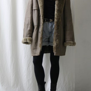 May include: A long, brown shearling coat with a fur collar and lining. The coat is worn over a black shirt and denim shorts. The person is wearing black tights and black ankle boots.