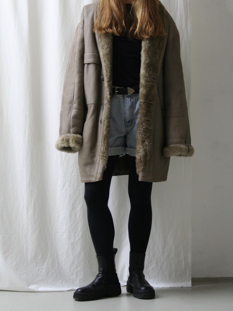 May include: A long, brown shearling coat with a fur collar and lining. The coat is worn over a black shirt and denim shorts. The person is wearing black tights and black ankle boots.