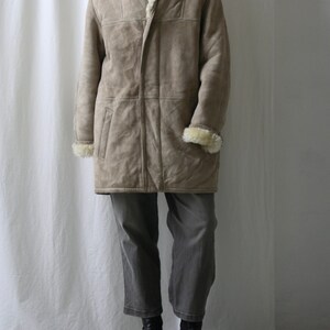 May include: A long, beige shearling coat with a fur-lined collar and a button closure. The coat is worn over gray pants and black boots.