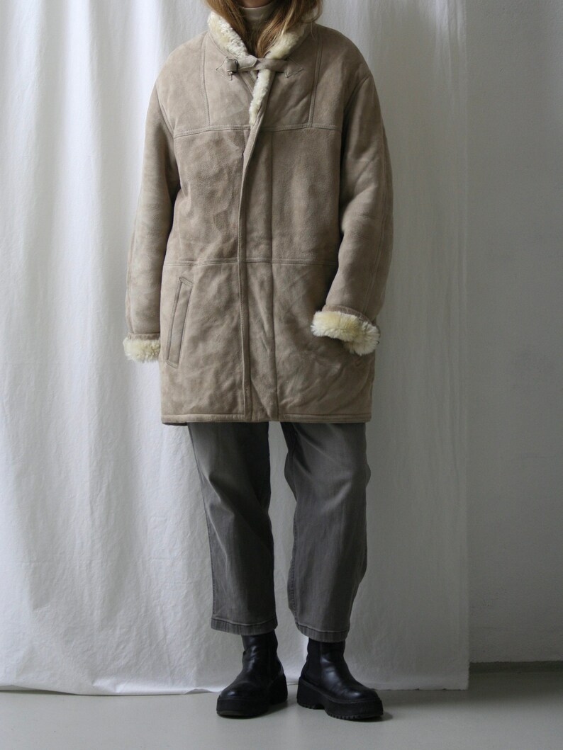 May include: A long, beige shearling coat with a fur-lined collar and a button closure. The coat is worn over gray pants and black boots.