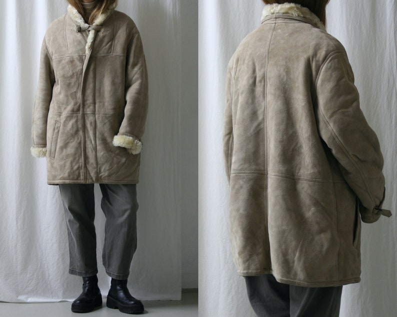 May include: A long, beige shearling coat with a fur collar and a zipper closure. The coat has a relaxed fit and is made of a soft, luxurious material.