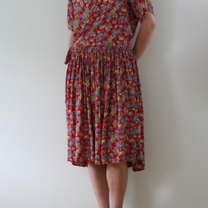 May include: A red floral print dress with a wrap-style bodice and a pleated skirt. The dress features short sleeves and a tie at the waist. The dress is knee-length and is paired with neutral-colored sandals.