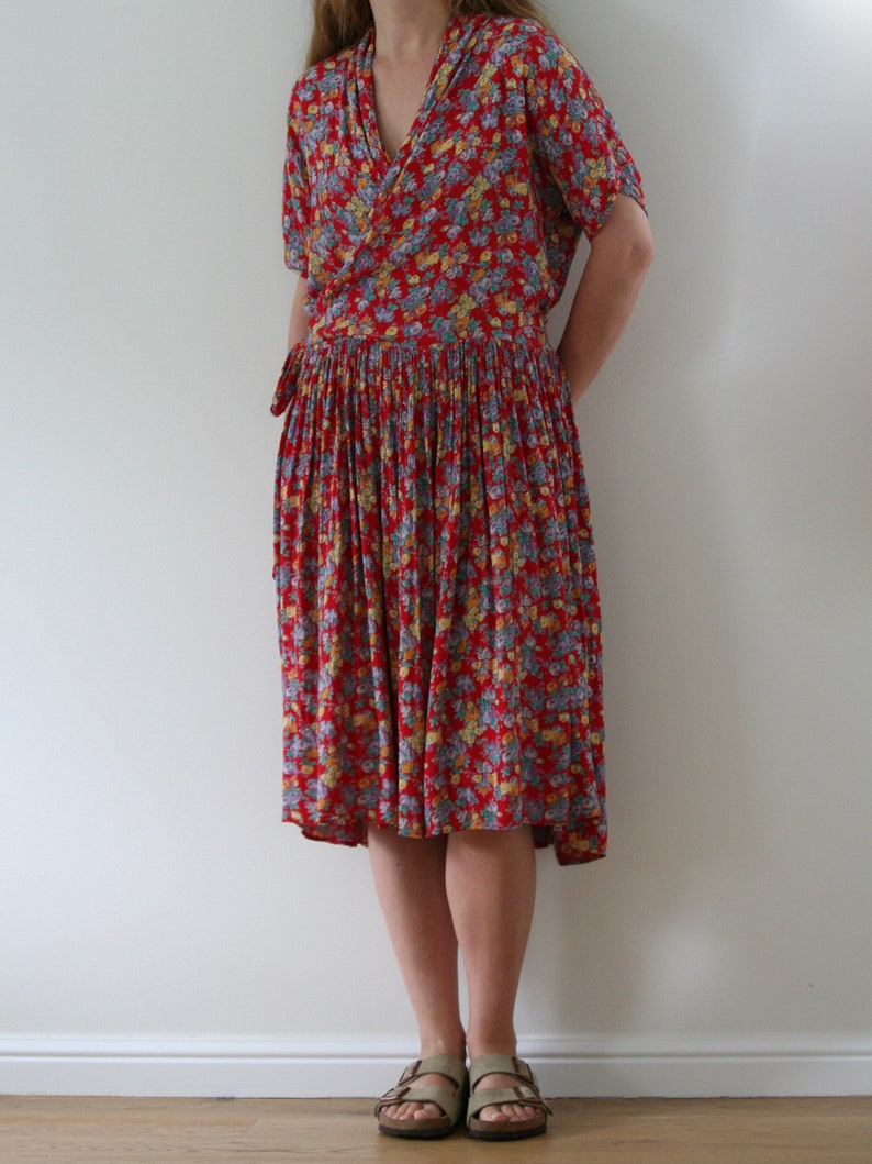 May include: A red floral print dress with a wrap-style bodice and a pleated skirt. The dress features short sleeves and a tie at the waist. The dress is knee-length and is paired with neutral-colored sandals.