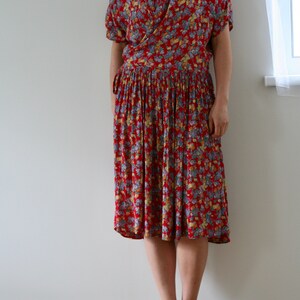 May include: A red floral print dress with short sleeves and a pleated skirt. The dress features a wrap-style bodice and a tie at the waist. The dress is knee-length and is paired with sandals.