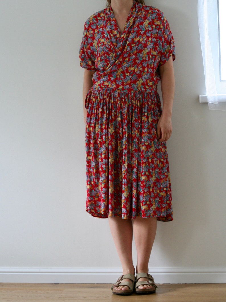 May include: A red floral print dress with short sleeves and a pleated skirt. The dress features a wrap-style bodice and a tie at the waist. The dress is knee-length and is paired with sandals.