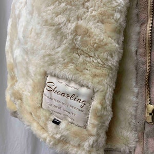 May include: A beige shearling jacket with a zipper closure. The jacket has a label that reads "The Original Shearling Guaranteed by Freetime The Best Quality Refinos Espanol 52 Made in Italy".