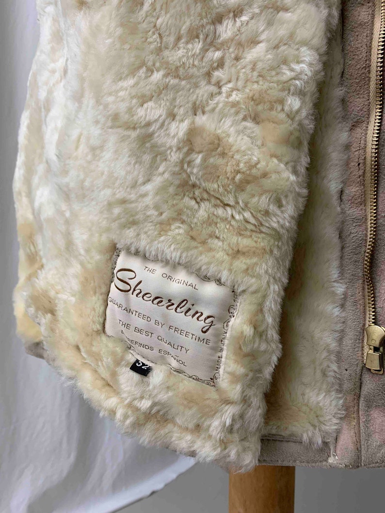 May include: A beige shearling jacket with a zipper closure. The jacket has a label that reads "The Original Shearling Guaranteed by Freetime The Best Quality Refinos Espanol 52 Made in Italy".