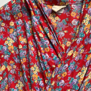 May include: Close-up of a red floral print garment. The fabric features a pattern of yellow, purple, and teal flowers. The garment has a wrap-style design with a collar and a textured surface. The overall aesthetic is vintage.