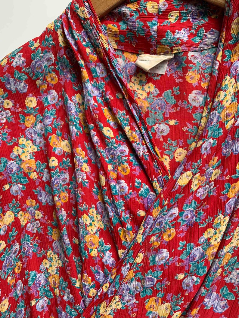 May include: Close-up of a red floral print garment. The fabric features a pattern of yellow, purple, and teal flowers. The garment has a wrap-style design with a collar and a textured surface. The overall aesthetic is vintage.