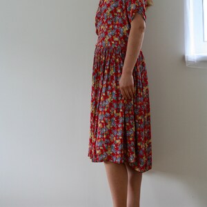 May include: A red floral print dress with short sleeves and a pleated skirt. The dress features a collared neckline and a fitted waist. The person is wearing light brown sandals with a cork sole.