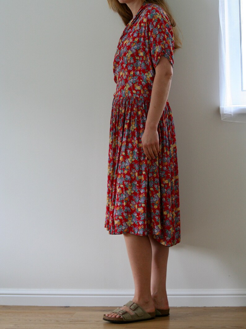 May include: A red floral print dress with short sleeves and a pleated skirt. The dress features a collared neckline and a fitted waist. The person is wearing light brown sandals with a cork sole.