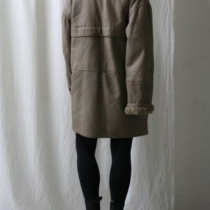 May include: A long, brown leather coat with a fur collar and a belt. The coat is worn by a person wearing black leggings and black boots.
