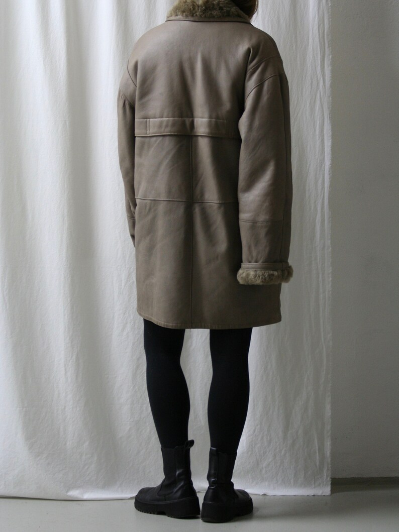 May include: A long, brown leather coat with a fur collar and a belt. The coat is worn by a person wearing black leggings and black boots.