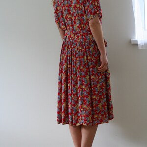 May include: A red floral print dress with short sleeves and a belted waist. The dress features a knee-length skirt and is adorned with a pattern of small flowers in shades of blue, yellow, and white. The dress is worn with sandals.