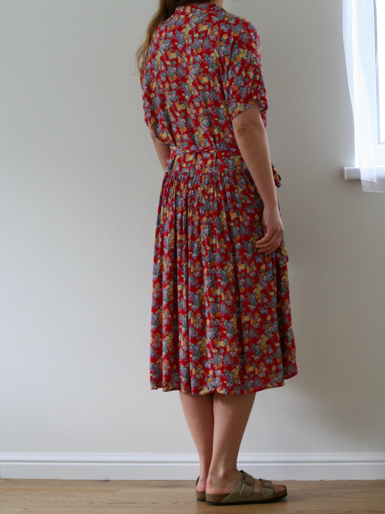 May include: A red floral print dress with short sleeves and a belted waist. The dress features a knee-length skirt and is adorned with a pattern of small flowers in shades of blue, yellow, and white. The dress is worn with sandals.
