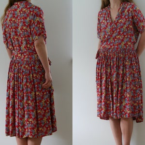 May include: A red floral print dress with short sleeves and a wrap-style bodice. The dress features a pleated skirt and a matching tie belt. The floral pattern includes yellow and blue accents. The dress falls to the mid-calf.