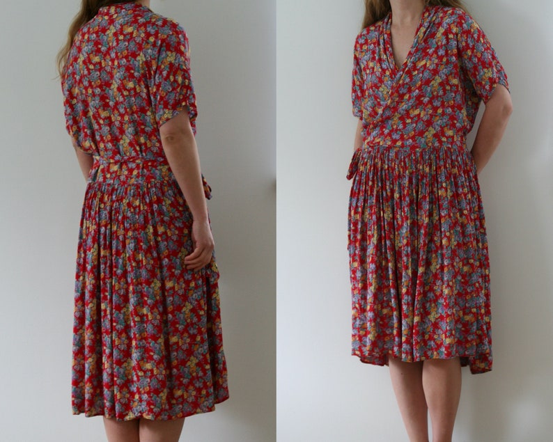 May include: A red floral print dress with short sleeves and a wrap-style bodice. The dress features a pleated skirt and a matching tie belt. The floral pattern includes yellow and blue accents. The dress falls to the mid-calf.