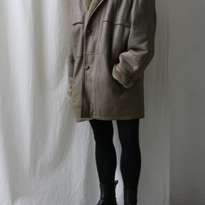 May include: A long, brown leather coat with a fur collar and lining. The coat has a button closure and two pockets. The model is wearing black tights and black ankle boots.