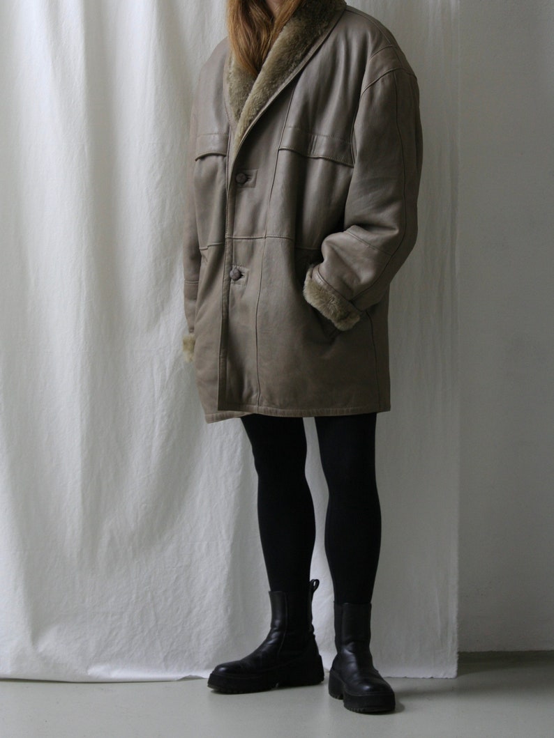 May include: A long, brown leather coat with a fur collar and lining. The coat has a button closure and two pockets. The model is wearing black tights and black ankle boots.