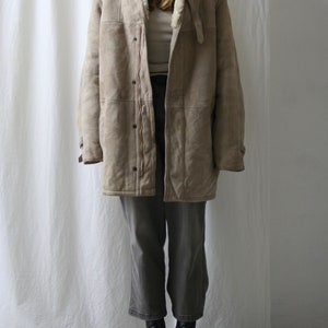 May include: A beige shearling coat with a white fur collar and a zipper closure. The coat is worn over a white turtleneck and gray pants.