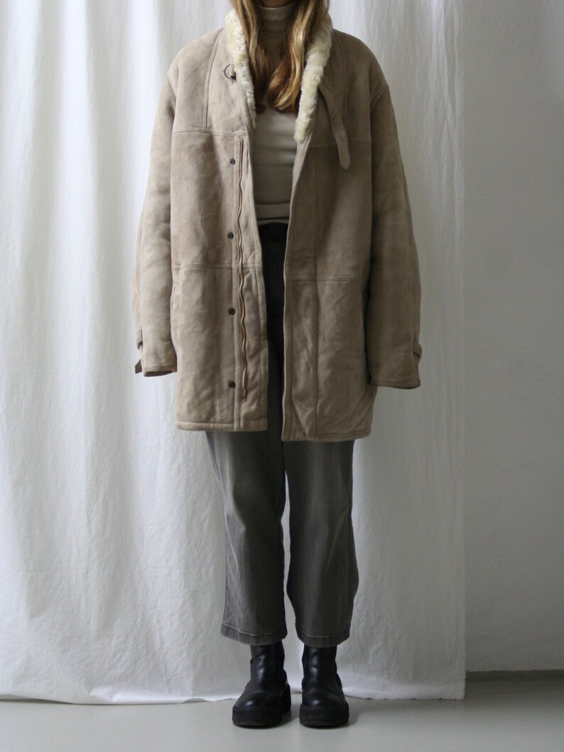 May include: A beige shearling coat with a white fur collar and a zipper closure. The coat is worn over a white turtleneck and gray pants.