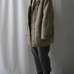 May include: A long, beige shearling coat with a fur collar and a button closure. The coat is worn over gray jeans and black boots.