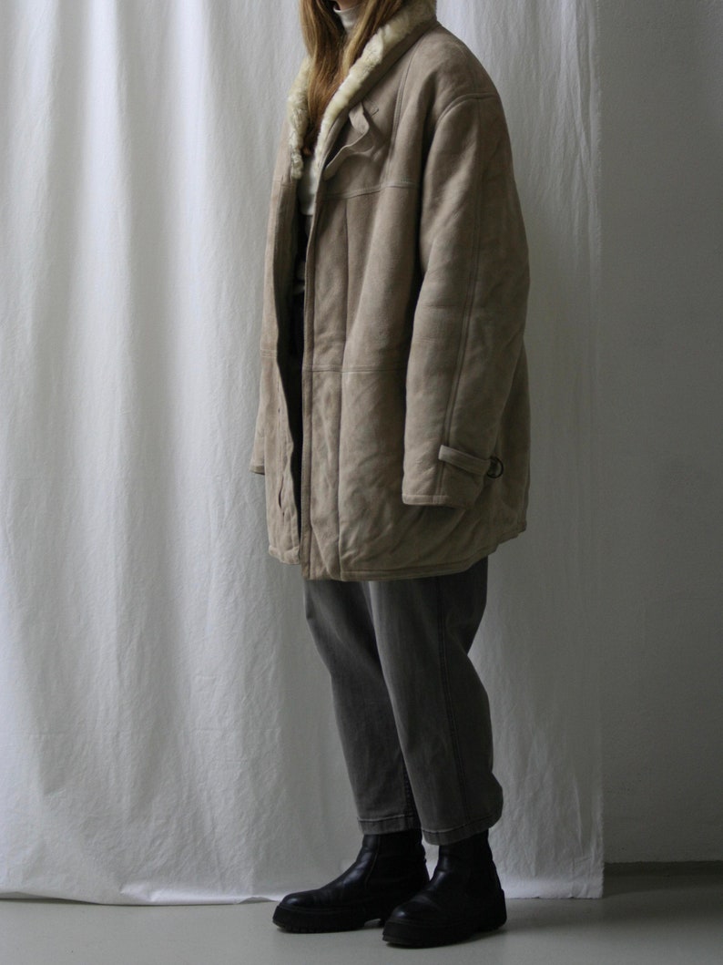 May include: A long, beige shearling coat with a fur collar and a button closure. The coat is worn over gray jeans and black boots.