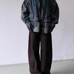 Könnte beinhalten: Eine Jeansjacke mit einzigartigem Design, die eine Mischung aus Texturen und Mustern aufweist. Die Jacke hat eine blau-graue Farbe mit schwarzen Akzenten und farbenfrohen Stoffdetails. Die Person trägt schwarze Handschuhe und eine braune Hose.