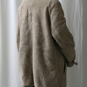 May include: A beige suede shearling coat with a fur collar. The coat has a button closure and a belt at the waist.