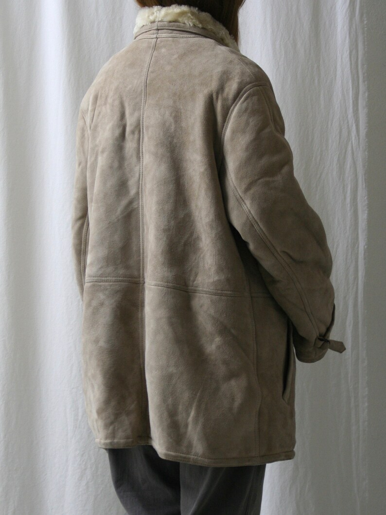 May include: A beige suede shearling coat with a fur collar. The coat has a button closure and a belt at the waist.