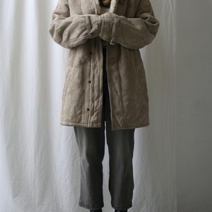 May include: A long, beige suede coat with a white faux fur collar and lining. The coat has a zipper closure and multiple pockets. The model is wearing the coat with grey pants and black boots.