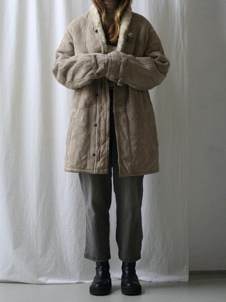 May include: A long, beige suede coat with a white faux fur collar and lining. The coat has a zipper closure and multiple pockets. The model is wearing the coat with grey pants and black boots.