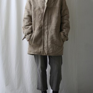 May include: A long, beige suede coat with a shearling collar and a button closure. The coat has a relaxed fit and is lined with a soft, white fleece.