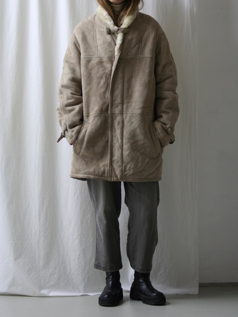 May include: A long, beige suede coat with a shearling collar and a button closure. The coat has a relaxed fit and is lined with a soft, white fleece.