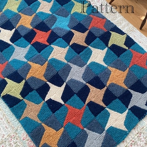 Op de afbeelding: Gebreide deken met een geometrisch sterrenpatroon in tinten blauw, oranje, beige en groen. De deken heeft een herhalend ontwerp van driedimensionale stervormen. De tekst "Knitting Pattern" is in witte letters geschreven.