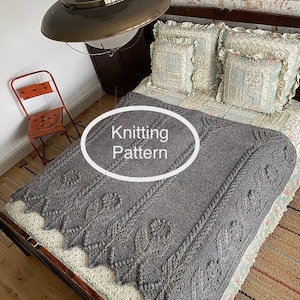 Może przedstawiać: Szary dzianinowy koc z wzorem warkoczowym i kwiatowym motywem. Koc leży na łóżku z poduszkami i drewnianym zagłówkiem. Tekst "Knitting Pattern" jest napisany na białym owalu.