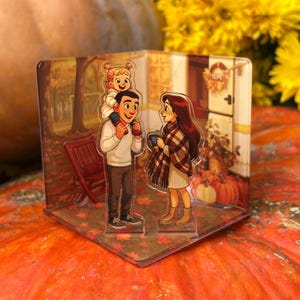 Puede incluir: Bloque acrílico transparente con una ilustración familiar. La obra de arte representa a un padre llevando a un niño sobre sus hombros y a una madre sosteniendo una taza. La escena está ambientada en un entorno otoñal con calabazas y follaje de otoño.