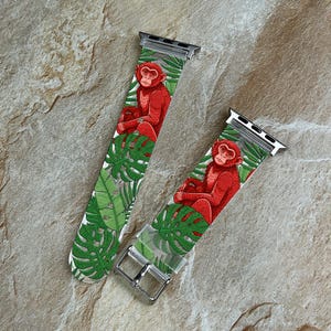 Può includere: Due cinturini per orologi con un design a scimmia rossa. I cinturini presentano una scimmia rossa seduta tra foglie tropicali verdi su sfondo bianco. I cinturini hanno connettori in metallo color argento e una fibbia argentata.