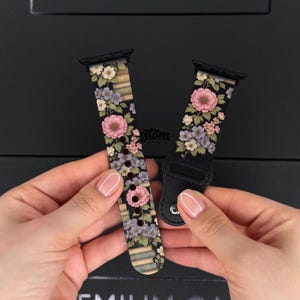 Puede incluir: Dos correas de reloj con estampado floral. Las correas presentan un fondo negro con flores rosas, moradas y blancas. Las correas están sujetas por las manos de una persona. Las correas son para un reloj inteligente.